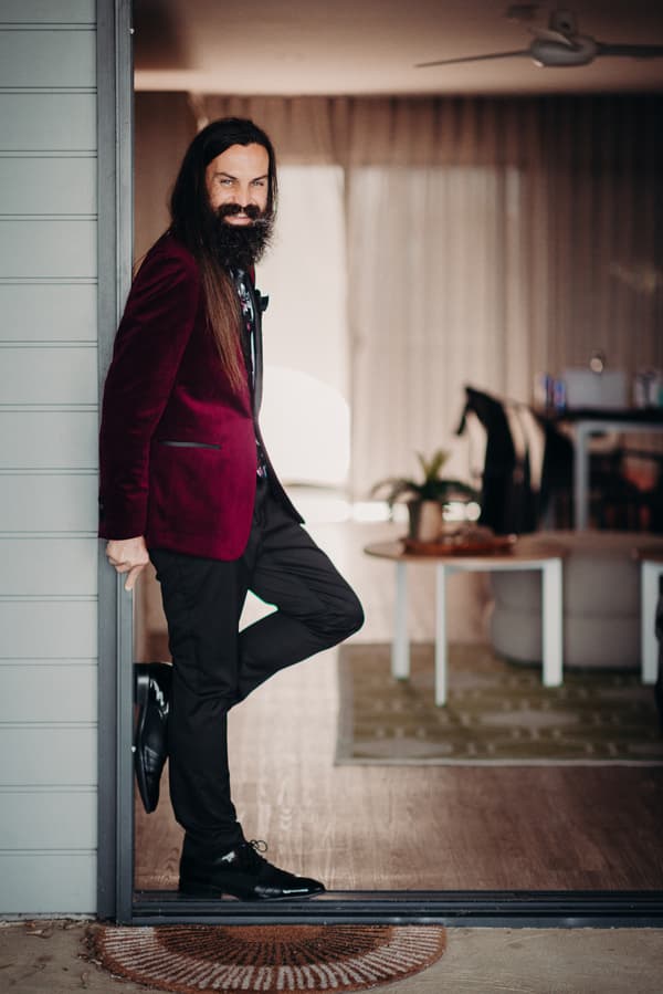 The groom David leans against a doorframe at Sandstone Point Hotel, wearing a burgundy jacket, black pants, and black shoes, indoors with a table and chairs visible in the background.