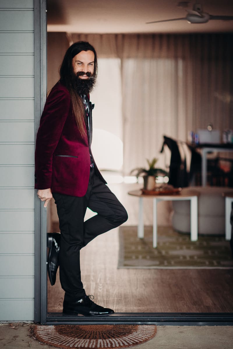 The groom David leans against a doorframe, wearing a burgundy jacket, black pants, and black shoes, indoors with a table and chairs visible in the background. Wedding live streaming by She Said Yes.