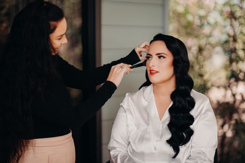 The bride Mindy sits wearing a white satin robe while a makeup artist applies makeup to her face at Sandstone Point Hotel.
