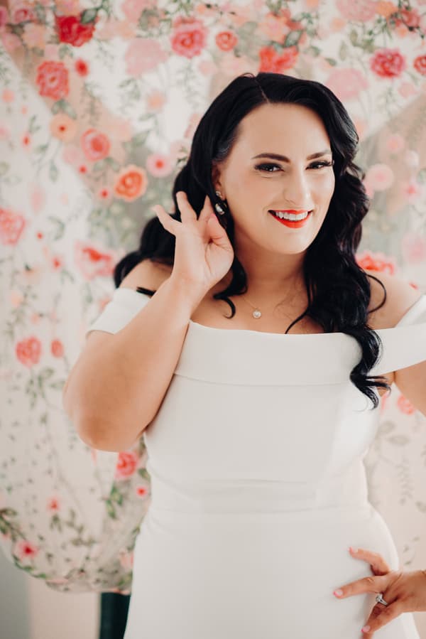 The bride Mindy poses in front of a floral backdrop wearing an off-shoulder white wedding dress, smiling and holding her hand near her ear.