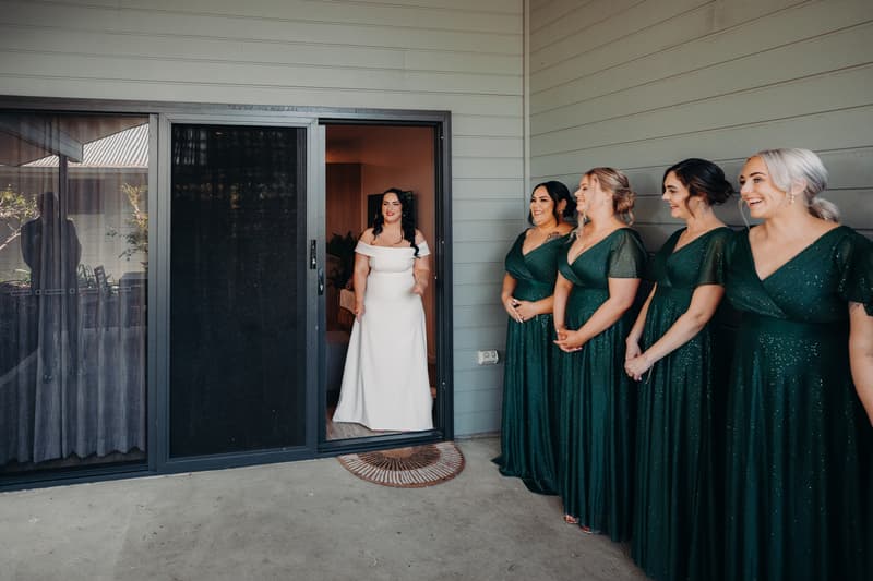 The bride Mindy stands in a doorway at Sandstone Point Hotel, smiling as four bridesmaids in matching green dresses stand in a row facing her.