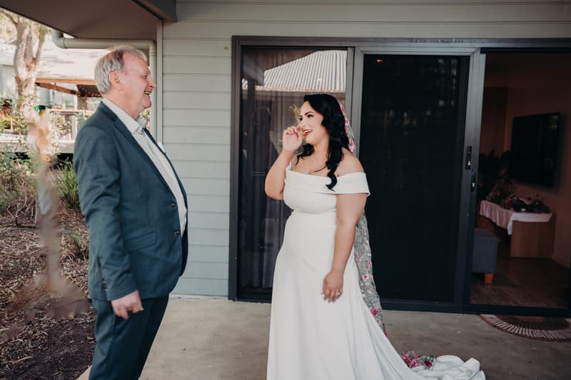The bride Mindy in a white off-shoulder wedding dress with a floral veil stands outside near a building, smiling and touching her face, while an older man, likely her father, in a suit smiles at her at Sandstone Point Hotel.