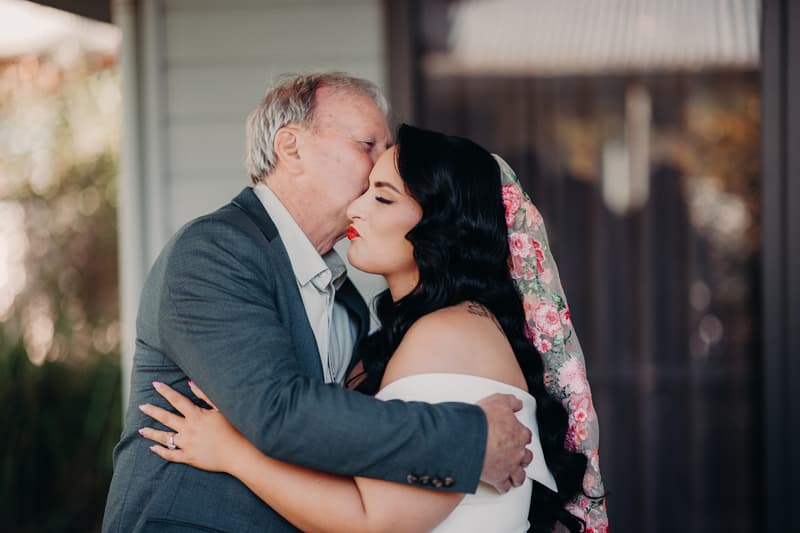 The bride Mindy is embraced and kissed on the forehead by an older man, likely her father, outside at Sandstone Point Hotel.