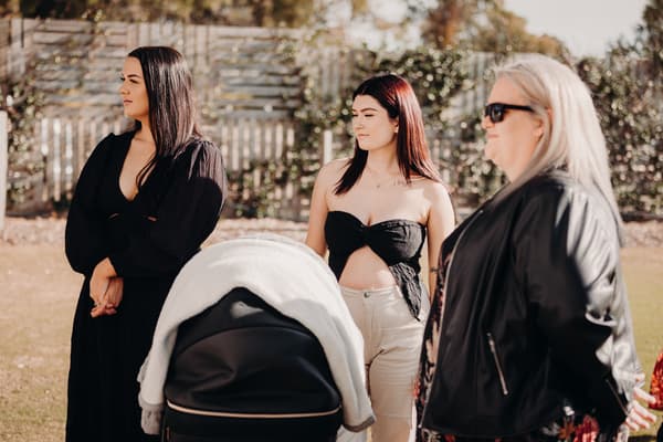 Three women stand outdoors near a stroller, with a wooden fence and greenery in the background. How much does a wedding photographer cost in Brisbane by She Said Yes.