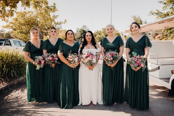 The bride Mindy stands with five bridesmaids dressed in matching dark green gowns, each holding bouquets of flowers, outside near a golf cart. Bridal editorial photography by She Said Yes.