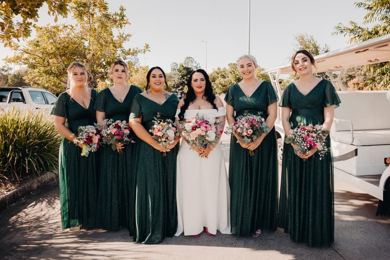 The bride Mindy stands with five bridesmaids dressed in matching dark green gowns, each holding bouquets of flowers, outside near a golf cart at Sandstone Point Hotel — Pavilion.