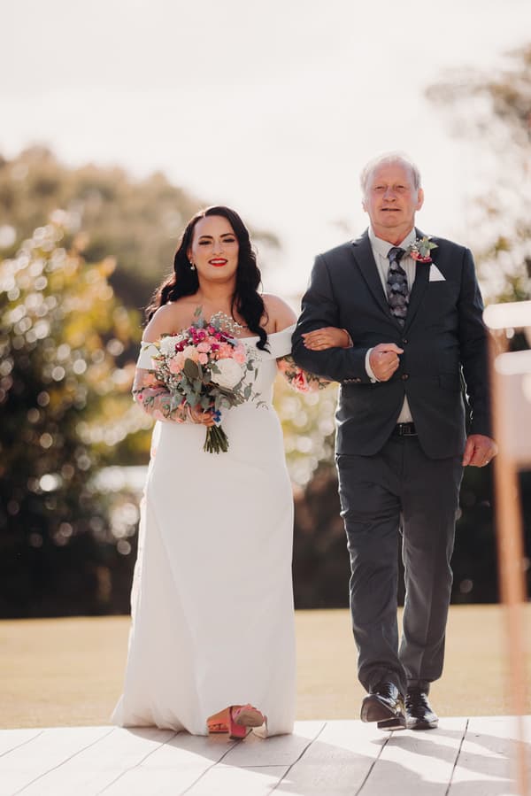 The bride walks arm-in-arm with an older man, likely her father, down an outdoor aisle at Sandstone Point Hotel — Pavilion, holding a bouquet of flowers.