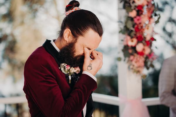 The groom David in a burgundy suit with a boutonniere, standing near a floral decorated pillar, touching his face with his eyes closed. Valentine's Day wedding photography by She Said Yes.