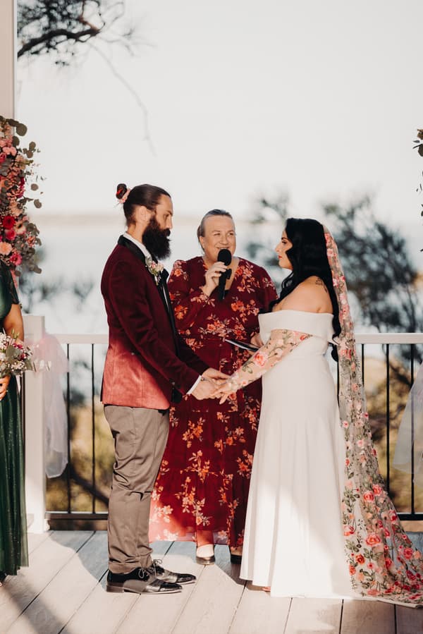 Bride Mindy and groom David hold hands facing each other while the officiant speaks into a microphone during their wedding ceremony at Sandstone Point Hotel — Pavilion.