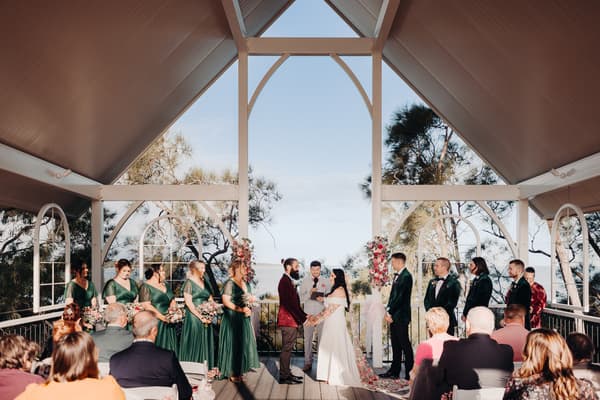 Bride Mindy and groom David hold hands facing each other during their wedding ceremony, with bridesmaids in green dresses and groomsmen in dark suits standing on either side, an officiant reading from a book, and guests seated watching. Australian wedding photographer by She Said Yes.