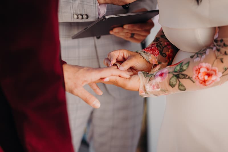 The bride places a wedding ring on the groom's finger during the ceremony at Sandstone Point Hotel — Pavilion, with the officiant holding a tablet in the background.