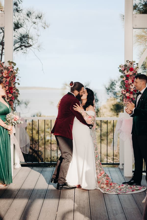 Bride Mindy and groom David share a kiss at the ceremony stage at Sandstone Point Hotel — Pavilion, flanked by a bridesmaid in a green dress holding a bouquet and a groomsman in a dark suit clapping.