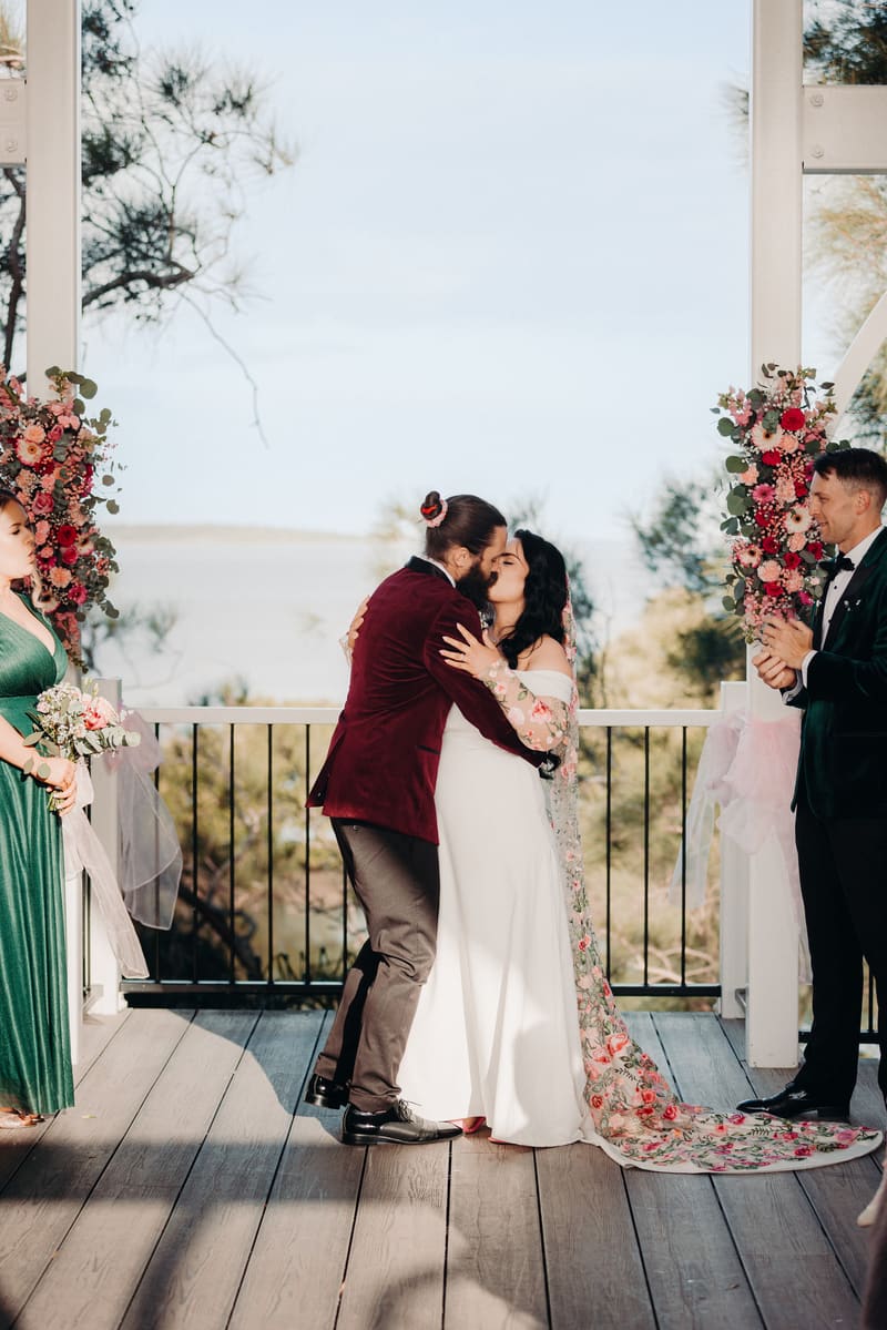 Bride Mindy and groom David share a kiss at the ceremony stage, flanked by a bridesmaid in a green dress holding a bouquet and a groomsman in a dark suit clapping. Brisbane videography by She Said Yes.