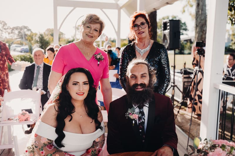 The bride Mindy and groom David sit at a table during their wedding ceremony at Sandstone Point Hotel — Pavilion, with two older women standing behind them and guests seated in the background.