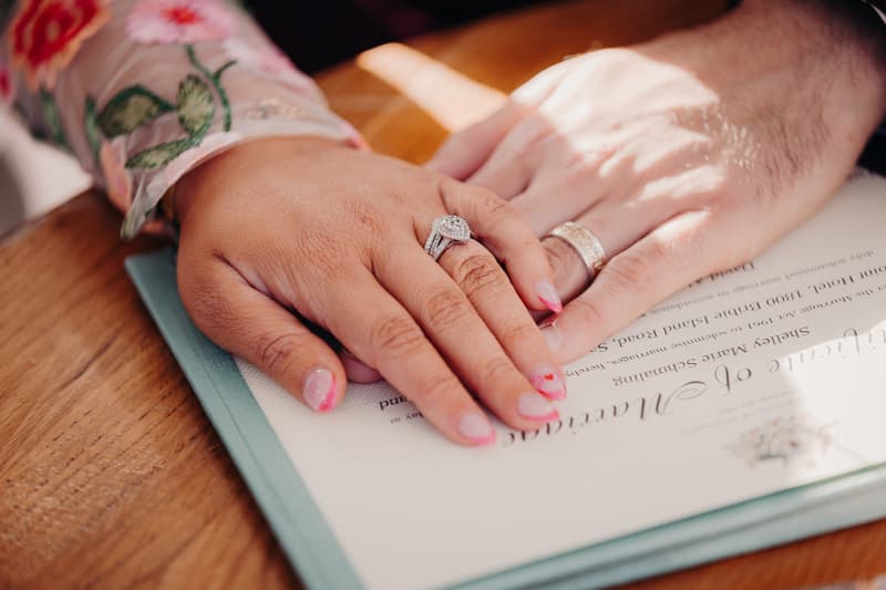 The bride and groom place their hands together on a wedding certificate at Sandstone Point Hotel — Pavilion, showing their wedding rings.