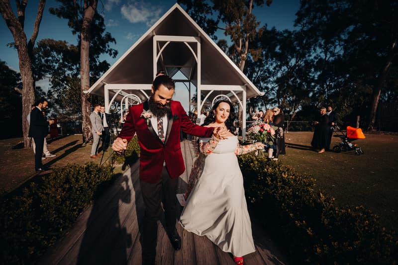 The bride Mindy and groom David walk hand in hand down the aisle at Sandstone Point Hotel — Pavilion after the ceremony, with guests standing and watching in the background.