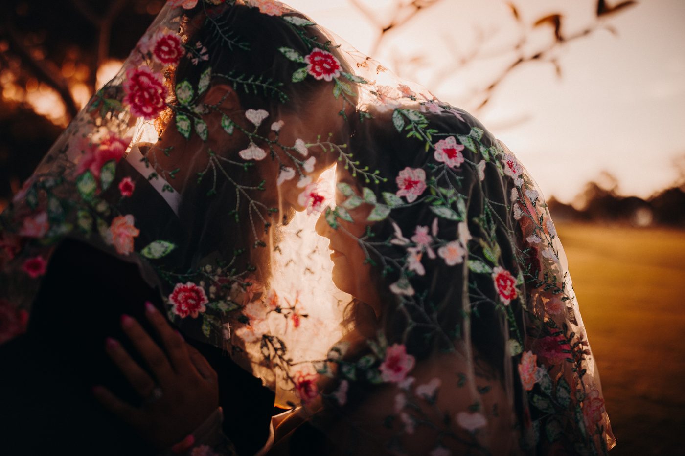 The bride Mindy and groom David stand close together under a floral embroidered veil during their couple portraits session at sunset. Cinematic videography wedding by She Said Yes.