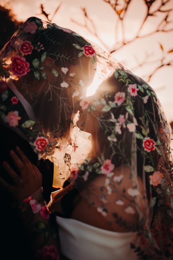The bride Mindy and groom David stand close together under a floral embroidered veil at Sandstone Point Hotel during their couple portraits session, with the sun setting behind them.