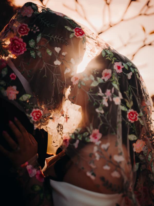 The bride Mindy and groom David stand close together under a floral embroidered veil at Sandstone Point Hotel during their couple portraits session, with the sun setting behind them.