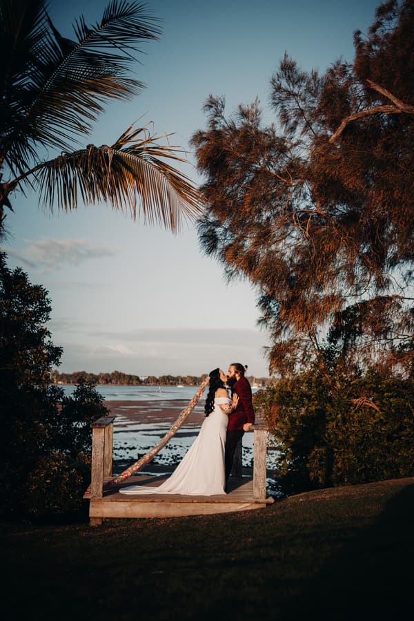 The bride and groom share a kiss on a small wooden platform overlooking a body of water at Sandstone Point Hotel, surrounded by trees and greenery.
