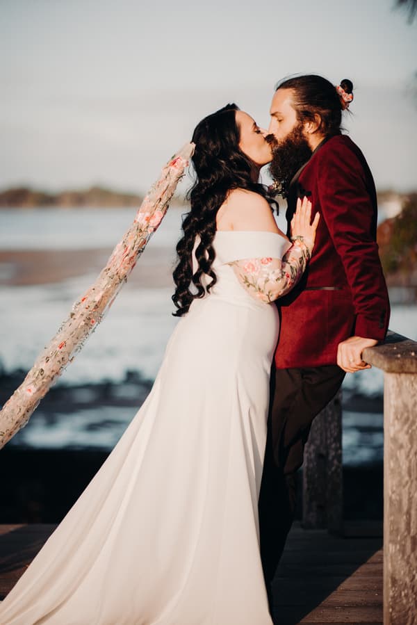 The bride Mindy in a white off-shoulder wedding gown with floral lace sleeves and veil kisses the groom David, who is wearing a burgundy jacket and black pants, at Sandstone Point Hotel near a waterfront.