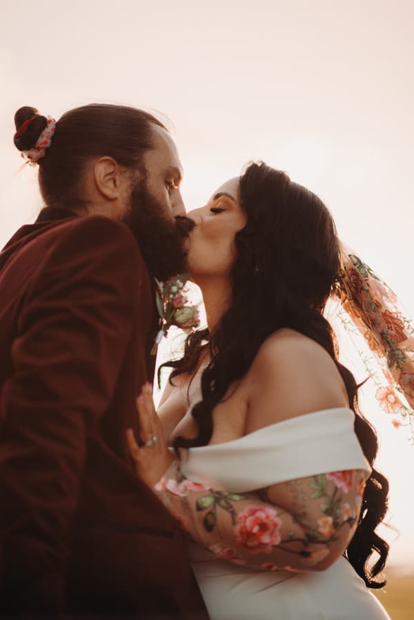 Bride Mindy and groom David share a kiss during their couple portraits session at Sandstone Point Hotel. Mindy wears an off-shoulder wedding dress with floral embroidered sheer sleeves and a matching floral veil, while David wears a dark suit with a boutonniere.
