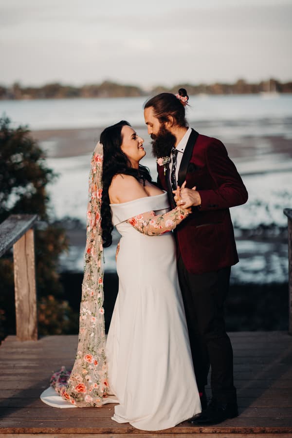 The bride Mindy and groom David pose together on a wooden deck at Sandstone Point Hotel, with water and distant trees in the background. Mindy wears an off-shoulder white wedding dress with floral lace sleeves and a long floral veil, while David wears a burgundy jacket with black lapels and a floral boutonniere.