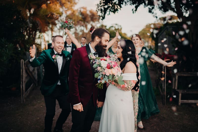 Bride Mindy and groom David stand close together holding hands, surrounded by bridesmaids and groomsmen celebrating behind them with flower petals falling. Photo and video packages by She Said Yes.