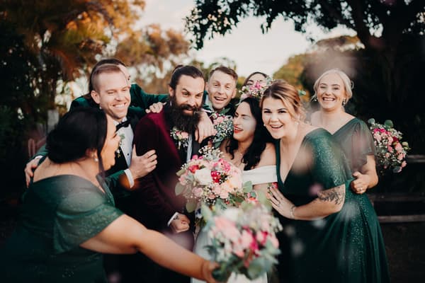 Mindy the bride and David the groom pose with bridesmaids and groomsmen outdoors, all smiling and holding bouquets. Best time of year to get married by She Said Yes.