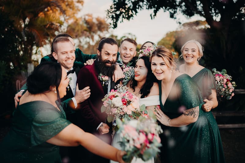 Mindy the bride and David the groom pose with bridesmaids and groomsmen outdoors, all smiling and holding bouquets. Best time of year to get married by She Said Yes.