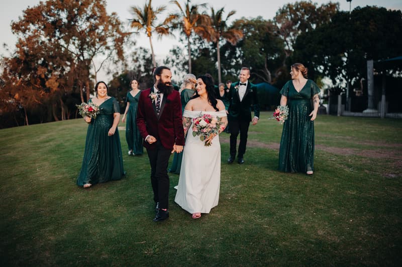 Mindy the bride and David the groom walk hand in hand on a grassy area at Sandstone Point Hotel, followed by bridesmaids in green dresses and groomsmen in dark suits.
