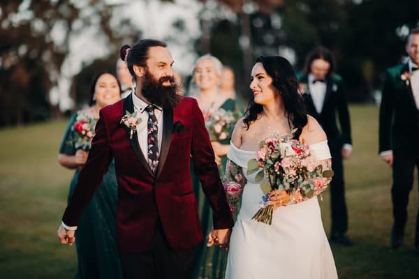 Bride Mindy and groom David walk hand in hand outdoors, followed by bridesmaids in green dresses and groomsmen in dark suits. Wedding photographers Brisbane by She Said Yes.