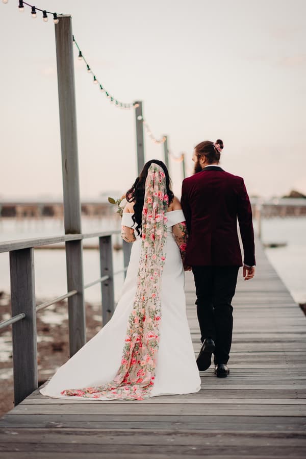 The bride Mindy and the groom David walk hand in hand along a wooden pier at Sandstone Point Hotel, with Mindy wearing a white off-shoulder gown and a long floral veil, and David in a dark burgundy jacket and black pants.