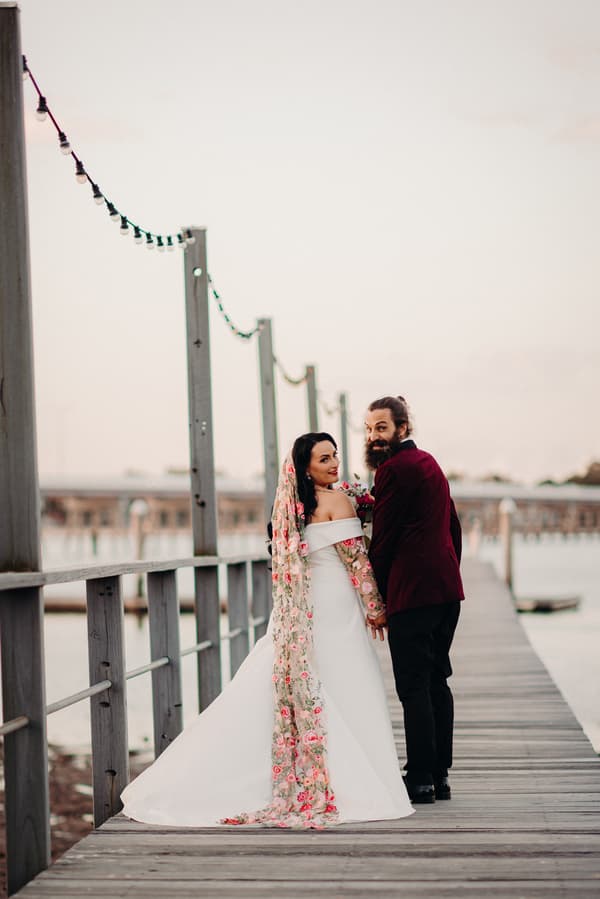 The bride Mindy in a white off-shoulder gown with a long floral veil and the groom David in a burgundy jacket and black pants stand holding hands on a wooden pier at Sandstone Point Hotel, looking back towards the camera.