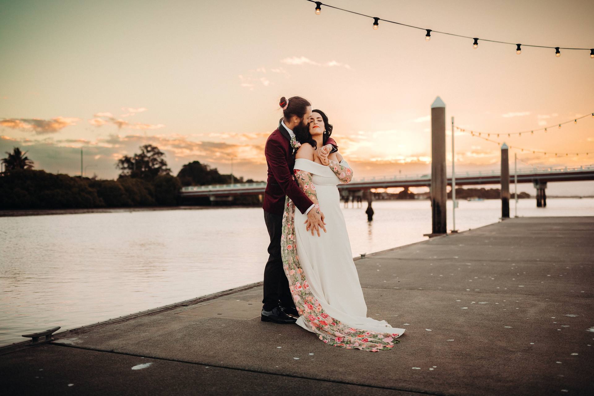 The bride and groom pose together on a waterfront pier at Sandstone Point Hotel during sunset, with the groom embracing the bride from behind.