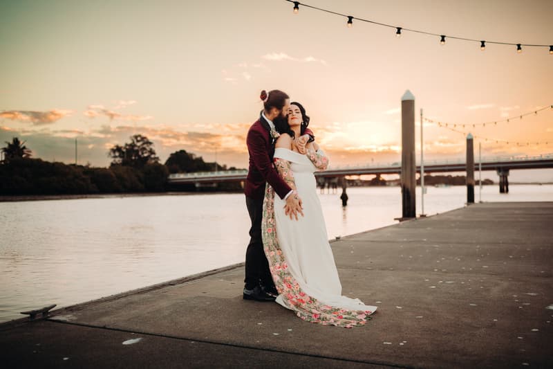The bride and groom pose together on a waterfront pier at Sandstone Point Hotel during sunset, with the groom embracing the bride from behind.
