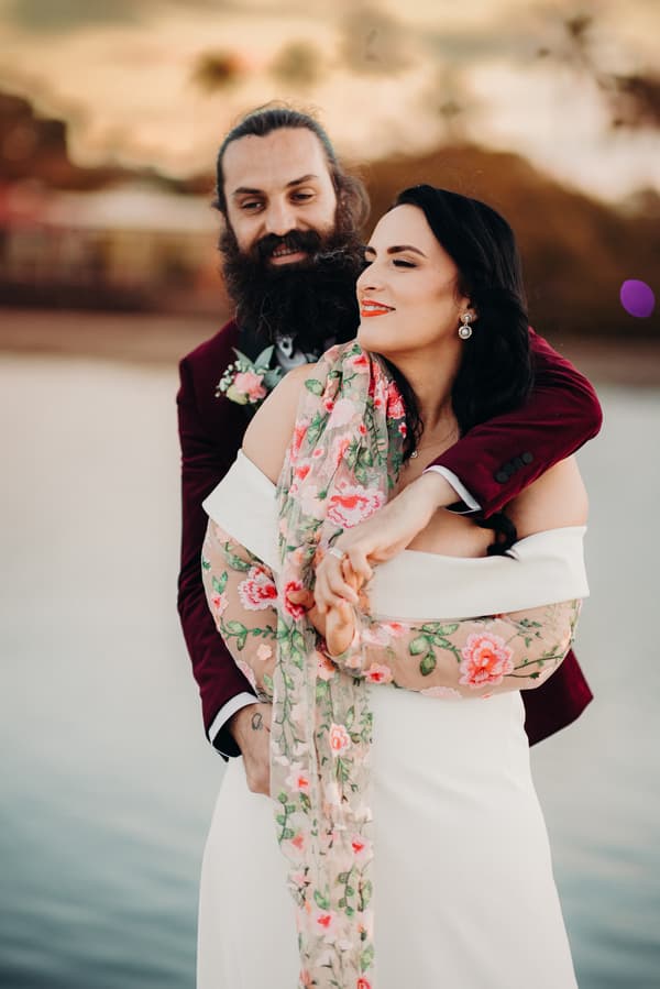 The bride Mindy and groom David embrace during their couple portraits session at Sandstone Point Hotel, with David wearing a burgundy suit and Mindy in a white dress with a floral embroidered shawl.
