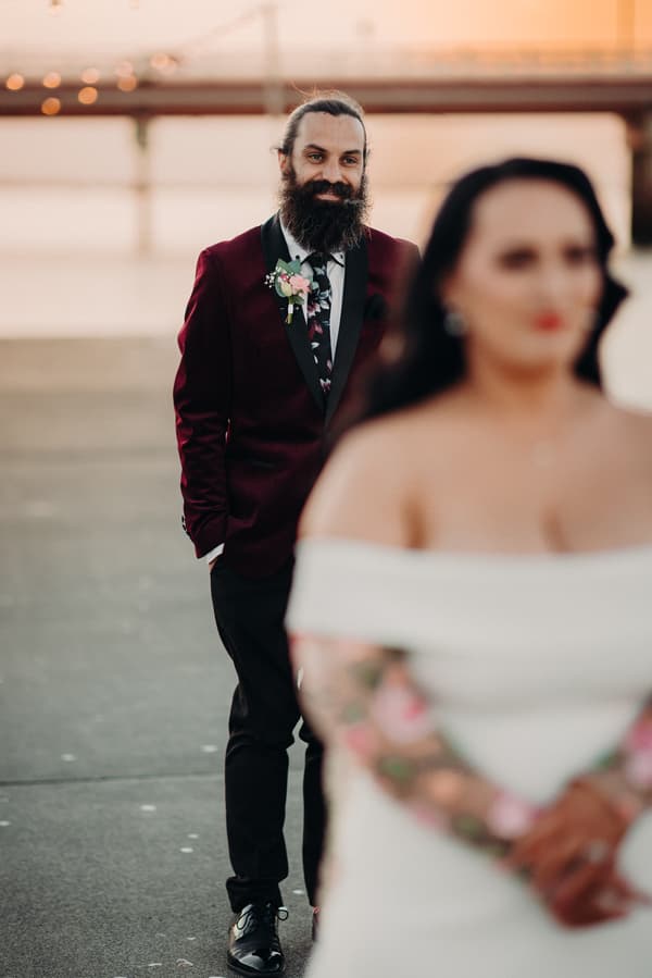 David, the groom, stands in a burgundy tuxedo jacket with a floral tie and boutonniere at Sandstone Point Hotel, with Mindy, the bride, in a white off-shoulder dress with floral tattoos on her arms blurred in the foreground.