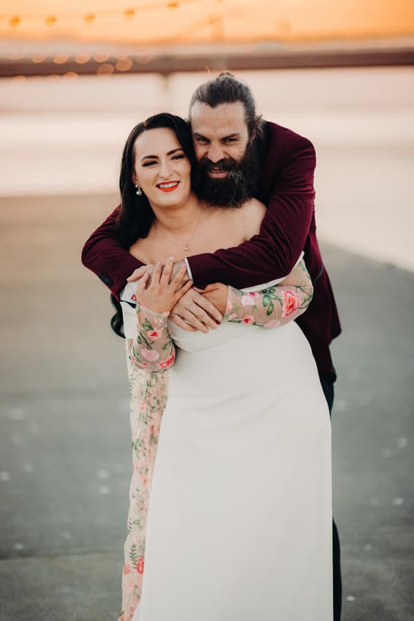 The bride Mindy in a white dress with floral sheer sleeves is embraced from behind by the groom David wearing a burgundy jacket at Sandstone Point Hotel during their couple portraits session.