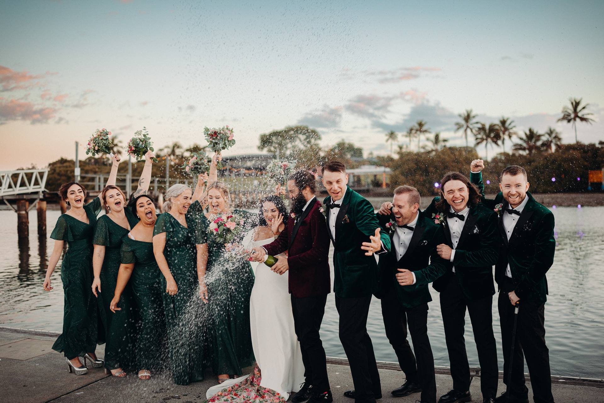 The bride Mindy in a white gown and the groom David in a dark suit celebrate with their bridesmaids in matching green dresses and groomsmen in dark suits by the water at Sandstone Point Hotel, as David sprays champagne.