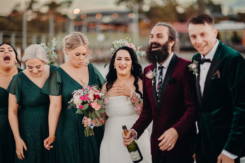 The bride Mindy in a white dress holding a bouquet stands with the groom David in a burgundy suit jacket holding a bottle, flanked by bridesmaids in dark green dresses and a groomsman in a dark green velvet jacket at Sandstone Point Hotel during couple portraits.