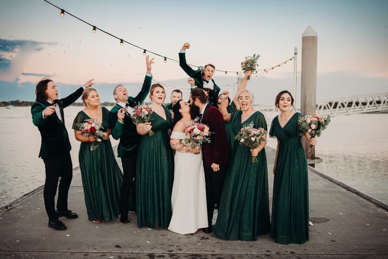 Bride Mindy and groom David kiss surrounded by bridesmaids in green dresses and groomsmen in dark suits on a pier at Sandstone Point Hotel with string lights overhead and water in the background.
