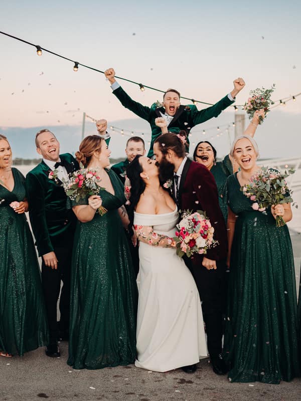 Mindy the bride and David the groom kiss surrounded by their bridal party on a pier at Sandstone Point Hotel, with bridesmaids in dark green dresses holding bouquets and groomsmen in dark suits celebrating.