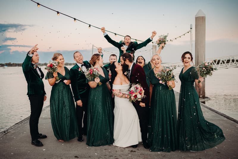 Mindy the bride and David the groom kiss surrounded by their bridal party on a pier at Sandstone Point Hotel, with bridesmaids in dark green dresses holding bouquets and groomsmen in dark suits celebrating.