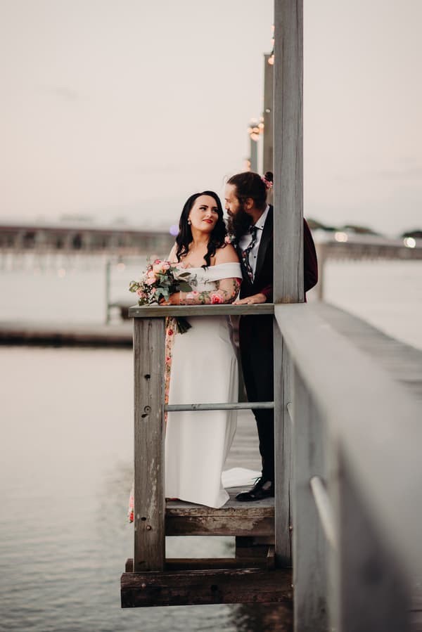 The bride Mindy and groom David stand together on a wooden balcony overlooking water at Sandstone Point Hotel, with Mindy holding a bouquet of flowers.