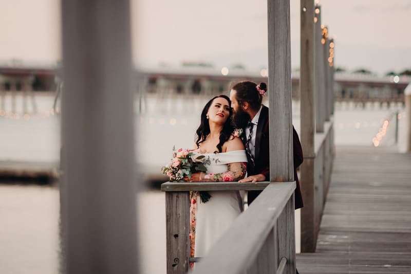 The bride Mindy in an off-shoulder white wedding dress holding a bouquet of flowers, and the groom David in a dark suit with a floral boutonniere, stand closely together on a wooden pier at Sandstone Point Hotel with string lights in the background.