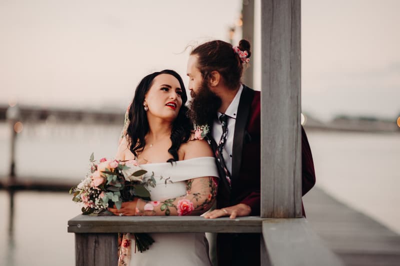 The bride Mindy in an off-shoulder white wedding dress holding a bouquet of flowers stands next to the groom David, who is wearing a dark suit with a floral tie, leaning on a wooden railing at Sandstone Point Hotel with a blurred waterfront background.