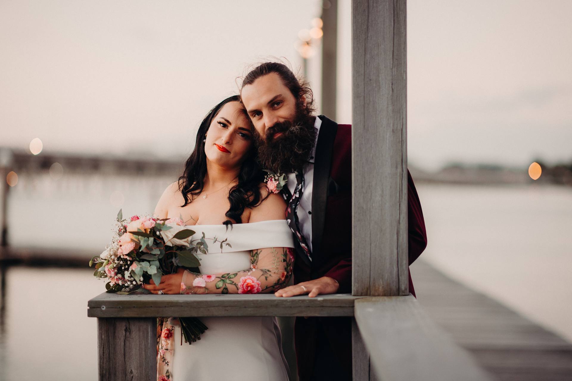 The bride Mindy in an off-shoulder white dress holding a bouquet of flowers leans against a wooden railing with the groom David in a dark suit standing close beside her at Sandstone Point Hotel.