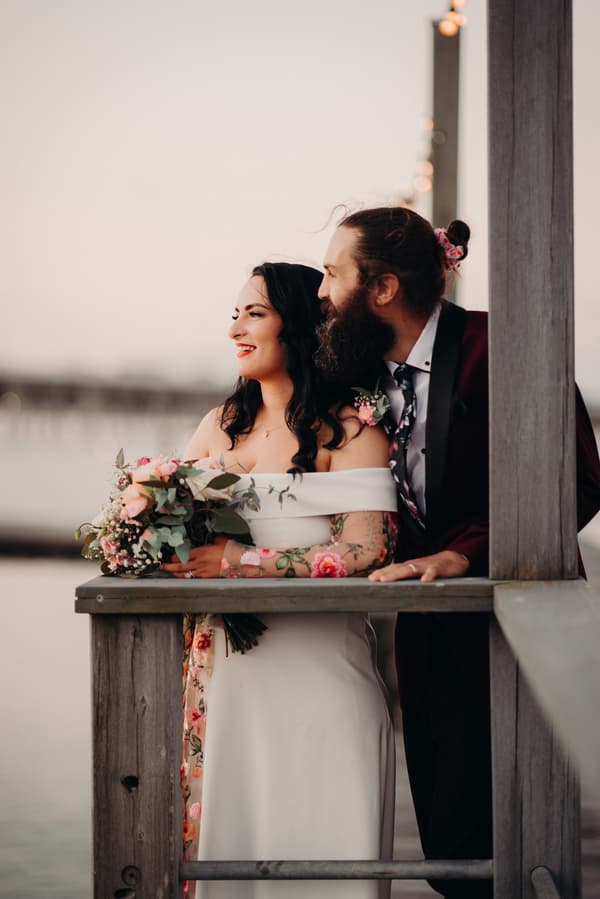 The bride Mindy in a white off-shoulder gown holding a bouquet of flowers stands next to the groom David, who is wearing a dark suit with a floral tie and boutonniere, leaning on a wooden railing at Sandstone Point Hotel during their couple portraits session.
