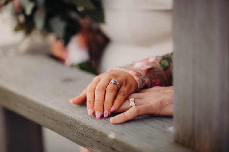 Close-up of the bride and groom's hands resting on a wooden surface at Sandstone Point Hotel, showing their wedding rings and the bride's floral sleeve.